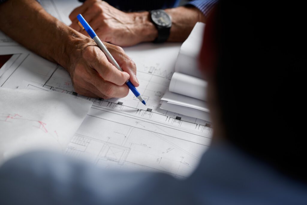 Close-up of an architect or engineer in Toronto reviewing and marking technical drawings with a pen, with rolled-up plans and a watch visible on the wrist.